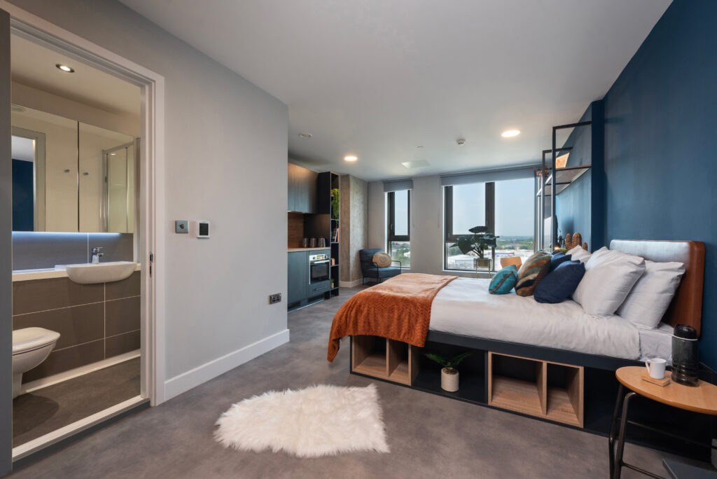 Luxury studio apartment at The Vantage in Nottingham, featuring modern furnishings, a large bed, desk area, kitchenette, and ample natural light through large windows.